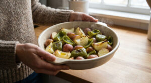 Woman's hands holding a steaming ceramic baking dish loaded with roasted vegetarian baked peppers and onions and red potatoes in a natural light kitchen setting.