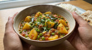 Woman holding a bowl of Aloo Hari Matar Foogath, a North Indian potato and green pea stew garnished with fresh cilantro.